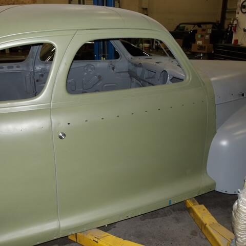 Classic car body in primer, undergoing restoration in a workshop.