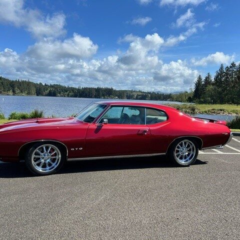 Red classic muscle car parked by a scenic lakeside under a blue sky.